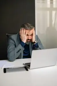A stressed adult man in a suit at an office desk with hands on face and laptop.